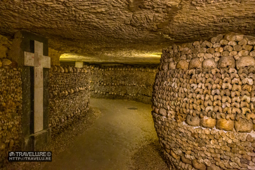 A massive barrel-shaped pillar supports the crossroads of underground aisles. On the left, you see a masonry monument - a tombstone with a cross. - the Catacombs in Paris - Travelure ©